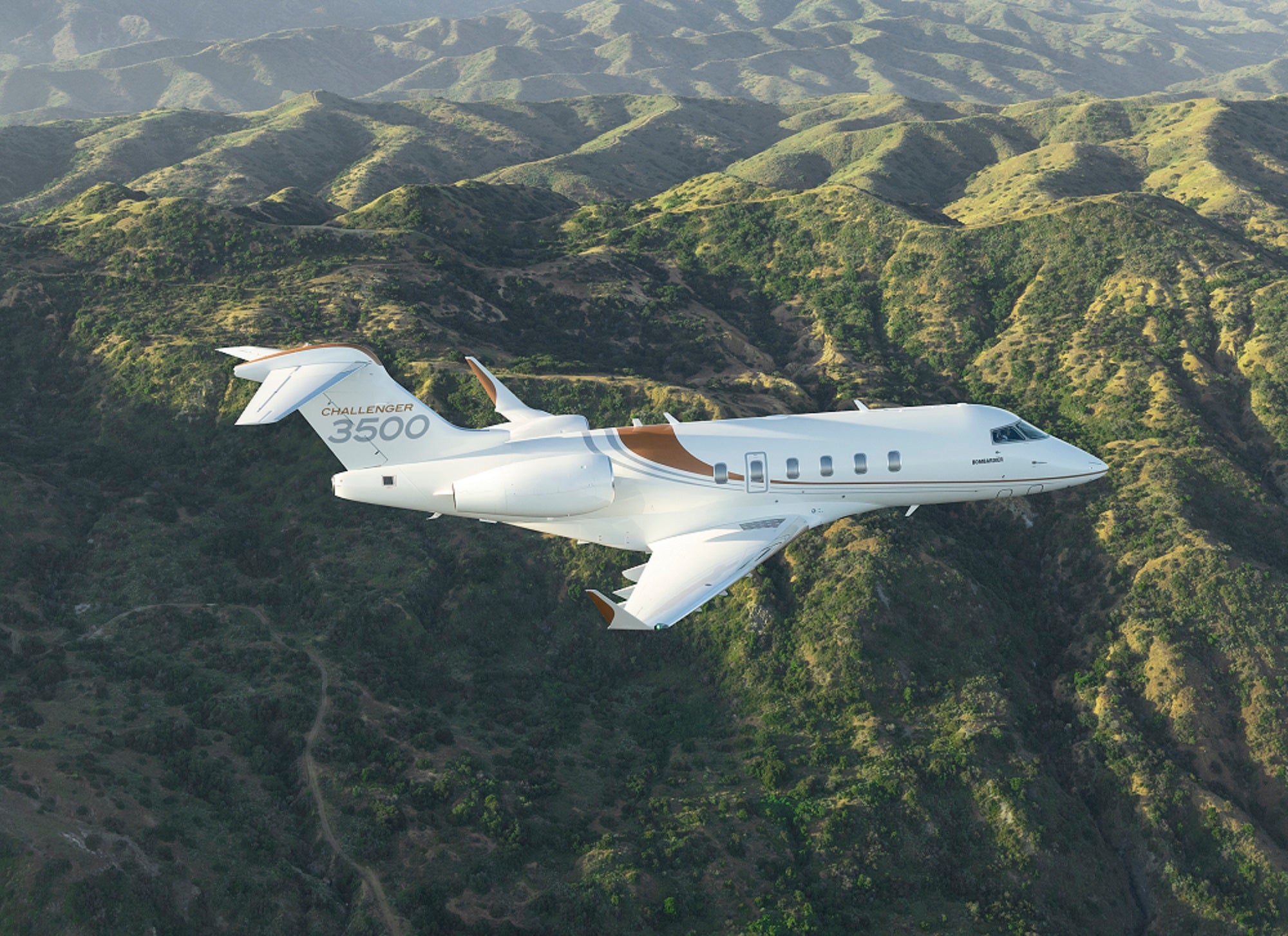 A Bombardier Challenger 3500 flies over a hilly, woodsy landscape.